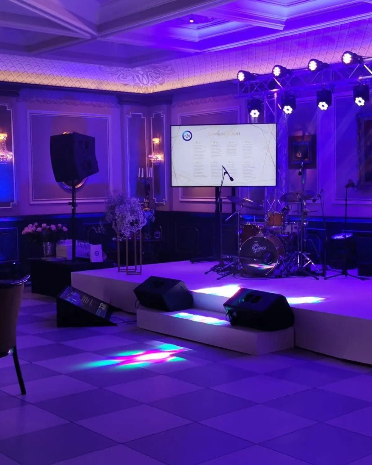 Scena su violetiniu apšvietimu, prožektoriais, ekranu ir muzikos instrumentais